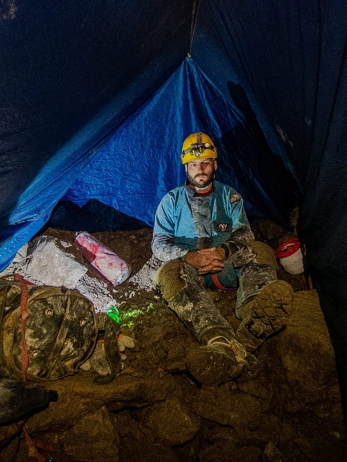 A caver protects himself from the cold wind and drips by staying under the tarp and behind a boulder while waiting for other cavers to climb the shaft.